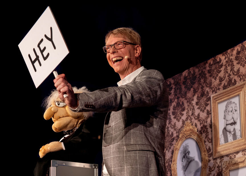 Bauchredner Jörg Jará zeigt Schild mit Aufdruck "hey" auf der Bühne
